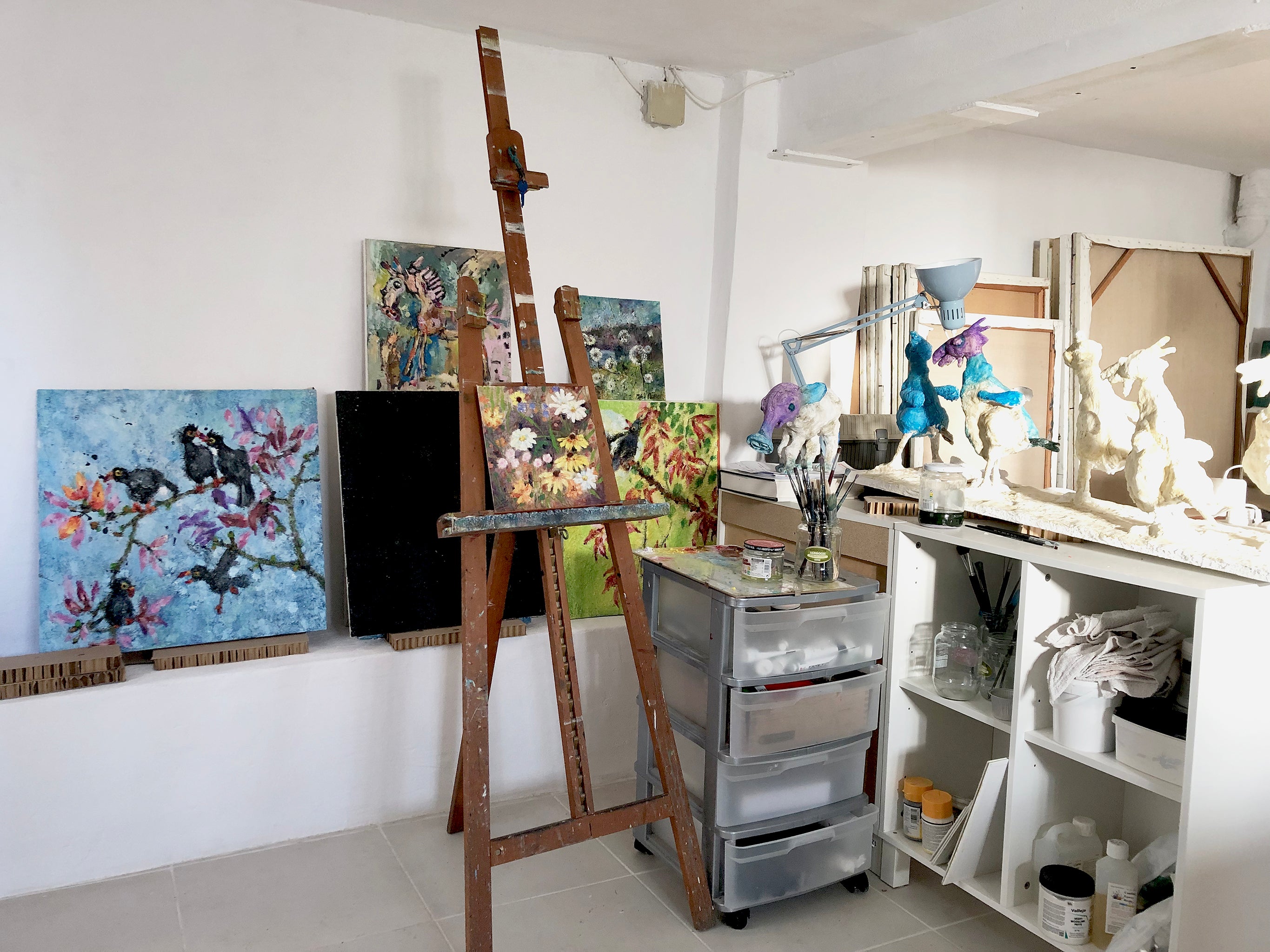 Art studio with easels, paintings, and sculptures on a white wall.